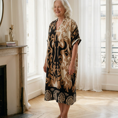 Femme de 70 ans portant la robe midi Célestine en Noir Absolu, imprimé ornemental noir et camel, dans un appartement parisien