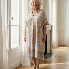 Femme de 70 ans portant la robe midi Célestine en Bleu Horizon, imprimé ornemental bleu clair et abricot, dans un appartement parisien