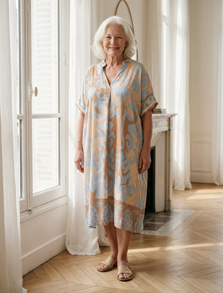 Femme de 70 ans portant la robe midi Célestine en Bleu Horizon, imprimé ornemental bleu clair et abricot, dans un appartement parisien